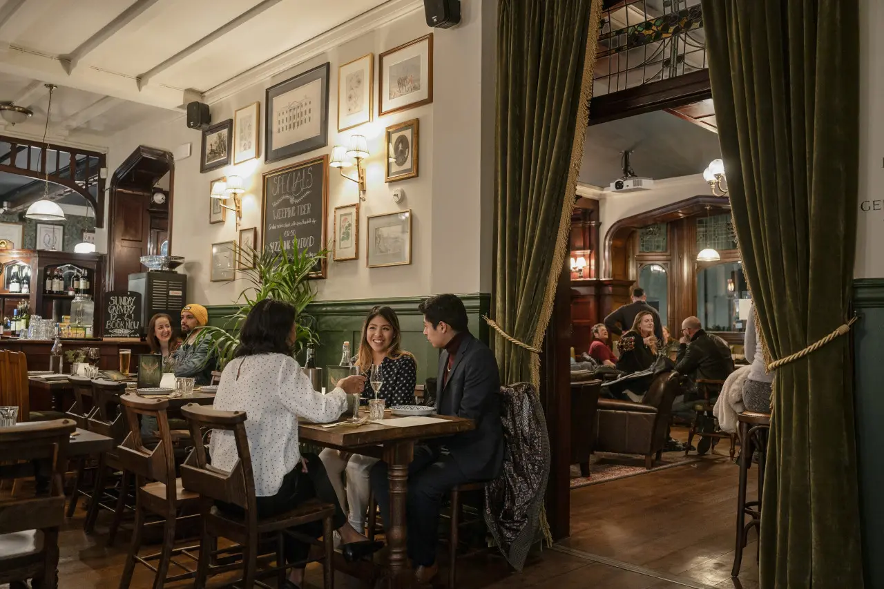 Photo of the interior of the Foerster by Fuller's. Groups of people are sat at tables. There are green curtains around a doorway which has art deco stained glass at the top. The walls are painted white with green wood panelling and have framed pictures on them